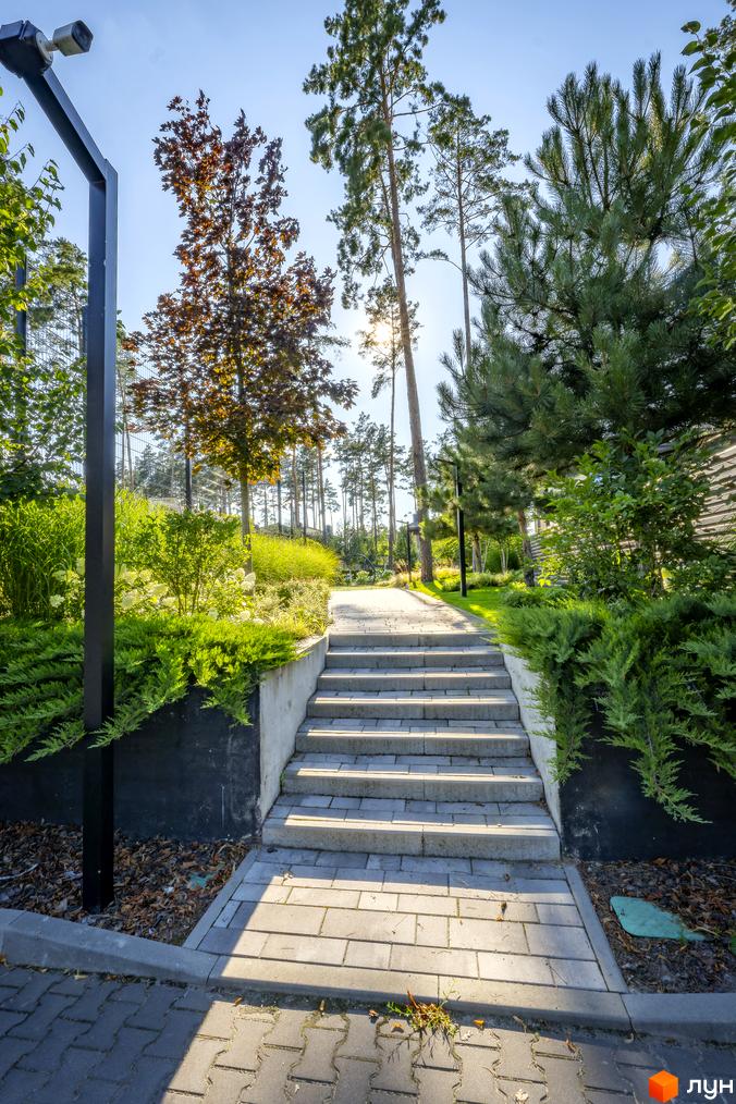 КМ ZAHRAVA village 2: кам'яні сходи ведуть до зеленої зони з деревами та кущами. Територія житлового комплексу доглянута та облаштована.