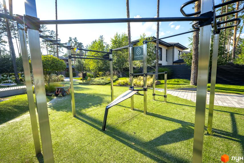 Зовнішній вигляд котеджного містечка ZAHRAVA village 2 з зоною для тренувань на відкритому повітрі. Навколо зелений газон, дерева та сучасна забудова.