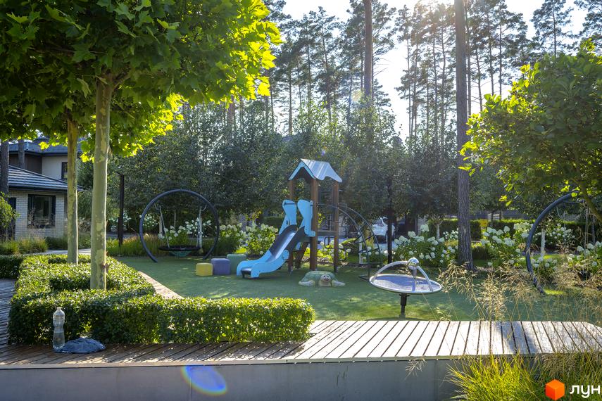 Дитячий майданчик у котеджному містечку КМ ZAHRAVA village 2 з гіркою, гойдалками та пісочницею. Навколо зелені дерева та кущі.