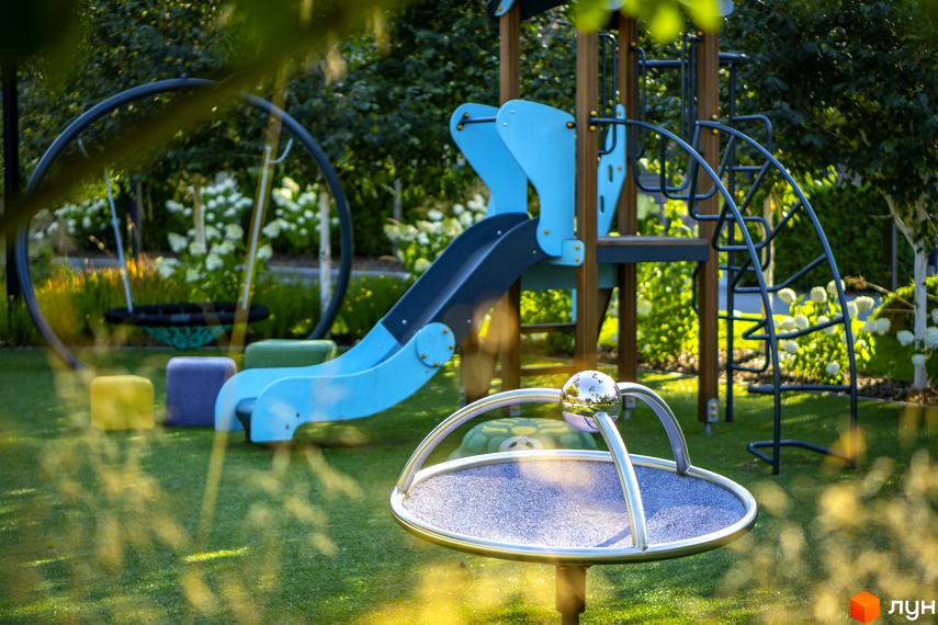 Дитячий майданчик у житловому комплексі КМ ZAHRAVA village 2 з гіркою, гойдалкою та каруселлю. Зелена галявина та квітучі рослини навколо.