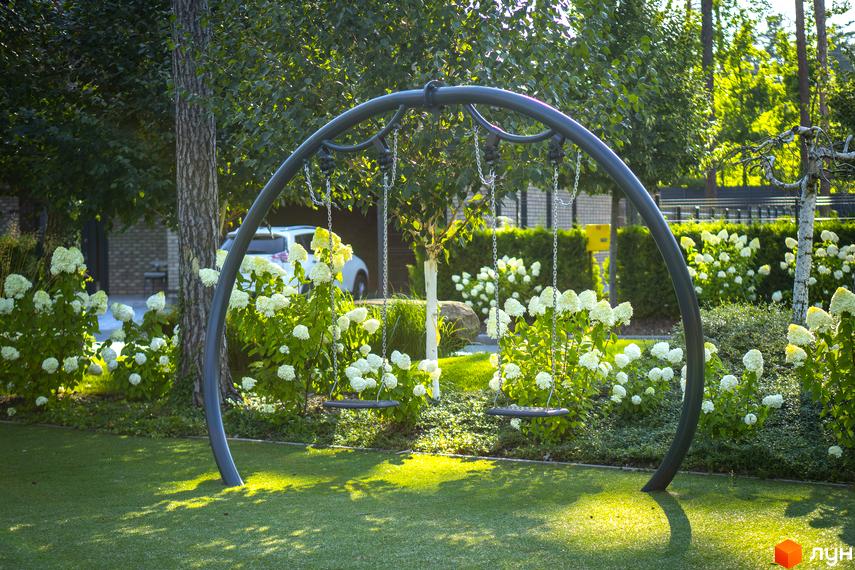 Котеджне містечко ZAHRAVA village 2: зелений двір з гойдалками та квітучими кущами. На задньому плані видно автомобіль та паркан.