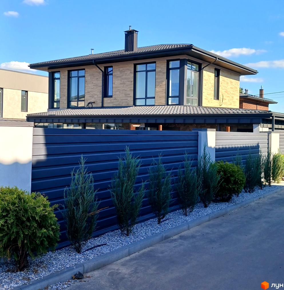 Зовнішній вигляд будинку з коричневою цегляною облицовкою та великими вікнами. Озеленена прибудинкова територія з парканом та туями.