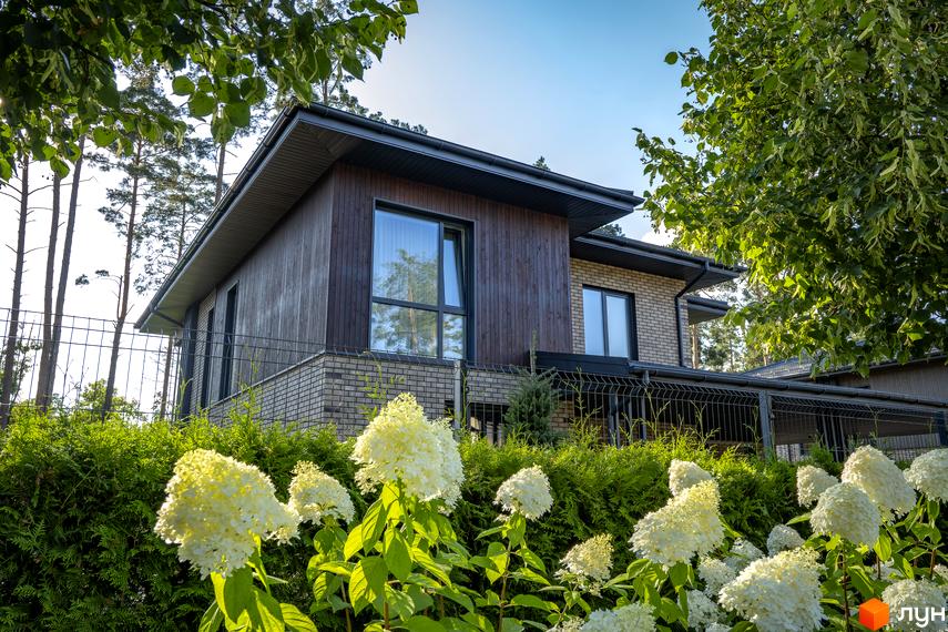 Котеджне містечко ZAHRAVA village 2: двоповерховий будинок з дерев'яним фасадом та цегляною обробкою, оточений зеленню та квітами.