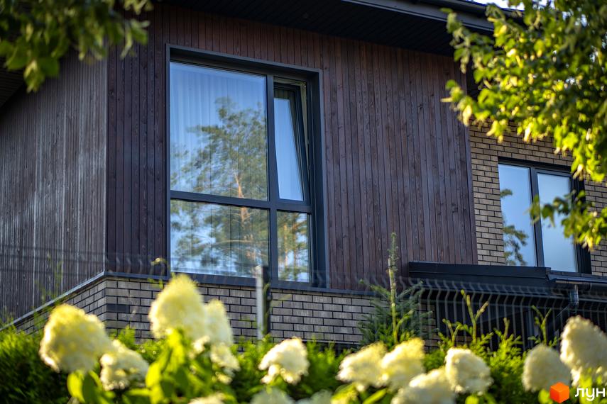 Котеджне містечко ZAHRAVA village 2: фасад будинку з дерев'яним оздобленням та цегляною кладкою, вікна з відображенням дерев. Прибудинкова територія з квітами та огорожею.