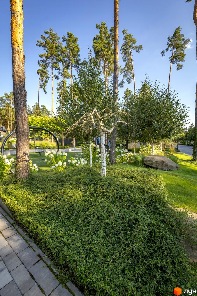 Котеджне містечко КМ ZAHRAVA village 2. Зелена прибудинкова територія з деревами, кущами та квітами, доріжка з бруківки та великий камінь.