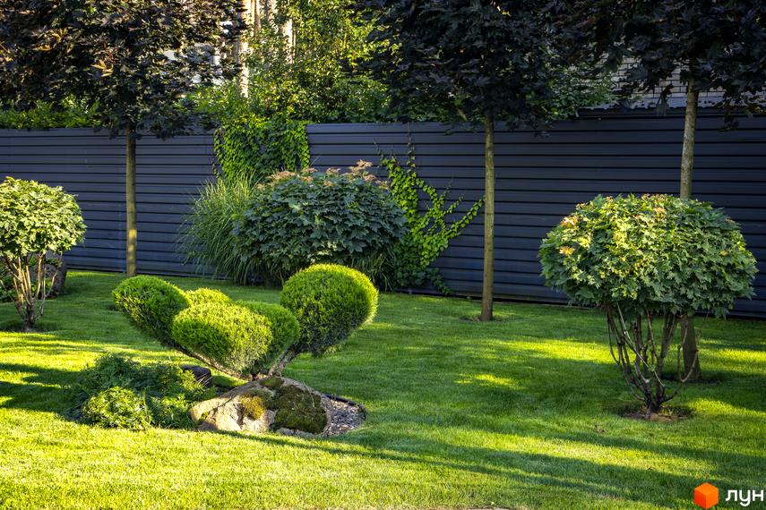 Доглянутий сад із акуратно підстриженими кущами та деревами, що оточують сучасний паркан. На передньому плані розташовані декоративні рослини та зелений газон.