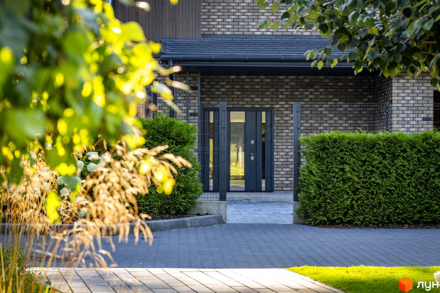 Котеджне містечко ZAHRAVA village 2. Вхідна група будинку з сірих цегляних стін, сірих дверей та металевого паркану. Прибудинкова територія з доглянутими кущами та бруківкою.