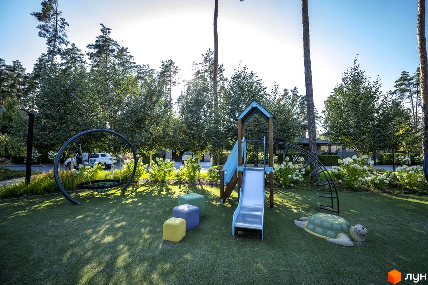 Дитячий майданчик у котеджному містечку КМ ZAHRAVA village 2 з гіркою, гойдалкою та ігровими елементами. Зелена територія з деревами та квітами.