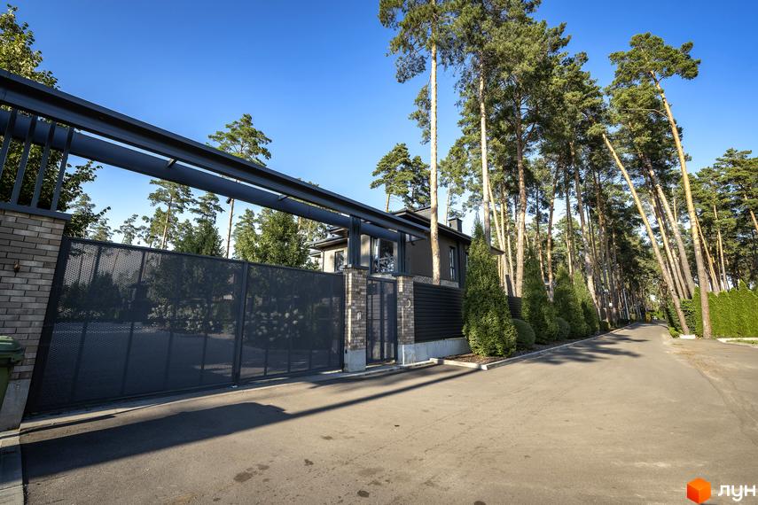 Котеджне містечко ZAHRAVA village 2. В'їзд на територію через сучасні ворота з металевою сіткою та цегляними колонами. Навколо високі сосни та зелені туї.