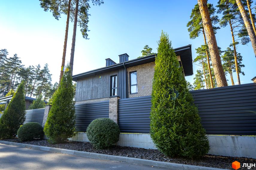 Котеджне містечко KМ ZAHRAVA village 2. Зовнішній вигляд будинку з цегляним фасадом та дерев'яними елементами, огороджений парканом. Прибудинкова територія з туями та кущами.