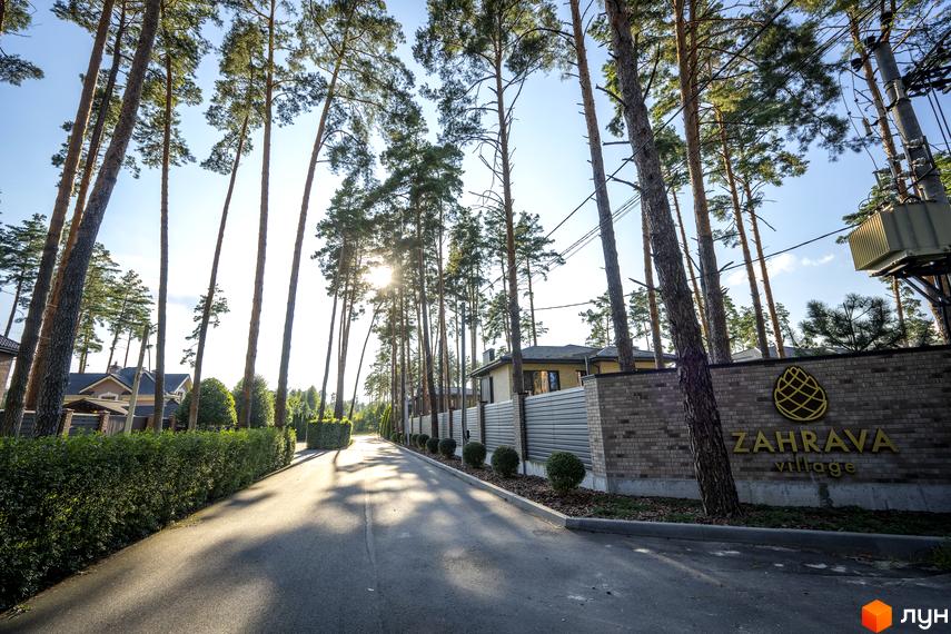 Котеджне містечко ZAHRAVA village з високими соснами та доглянутими зеленими насадженнями вздовж дороги. На фасаді видно цегляну огорожу з логотипом та назвою комплексу.