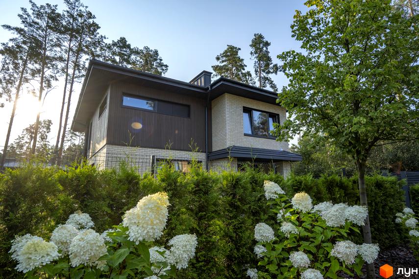 Котеджне містечко КМ ZAHRAVA village 2: двоповерховий будинок з цегляним та дерев'яним фасадом, вікнами та дахом темних кольорів, оточений зеленою огорожею та квітучими рослинами.