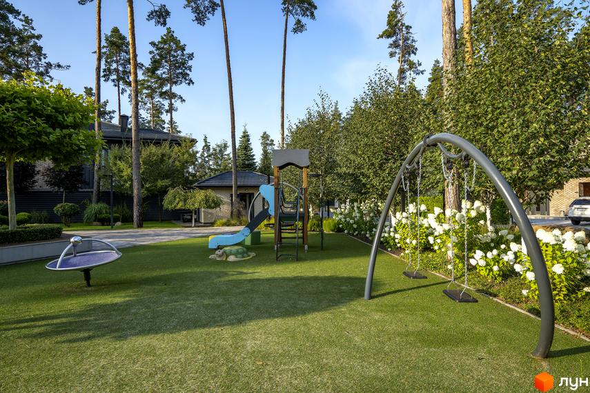 Дитячий майданчик з гойдалками та гіркою на території котеджного містечка КМ ZAHRAVA village 2. Навколо зелені насадження та сучасні будинки.