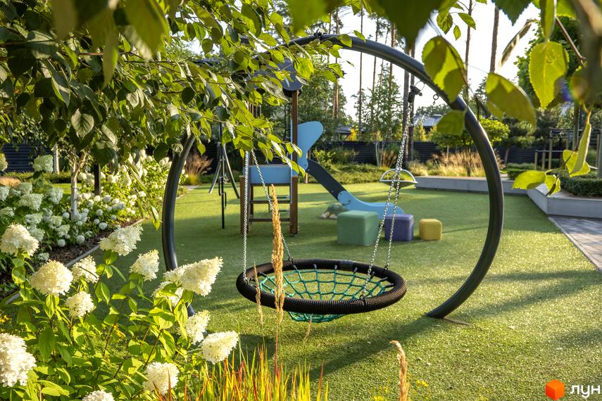 Дитячий майданчик з гойдалкою-гніздом та гіркою у котеджному містечку КМ ZAHRAVA village 2. Навколо зелена галявина та квітучі кущі.