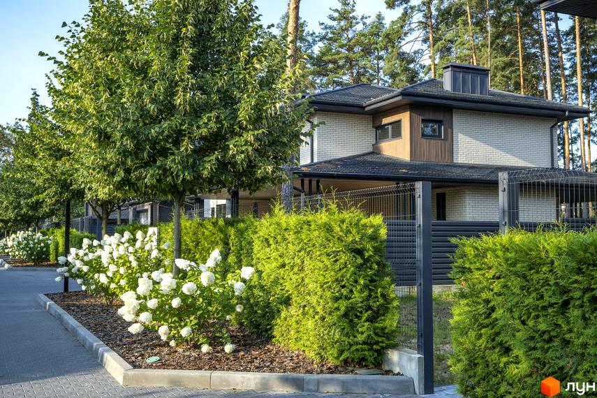 Котеджне містечко ZAHRAVA village 2 з білою цегляною фасадною частиною та зеленими туями. Навколо будинку облаштована прибудинкова територія з деревами та квітами.