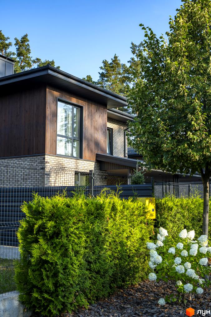 Котеджне містечко ZAHRAVA village 2: двоповерховий будинок з дерев'яним фасадом та цегляною кладкою, зелені туї та білі квіти на прибудинковій території.