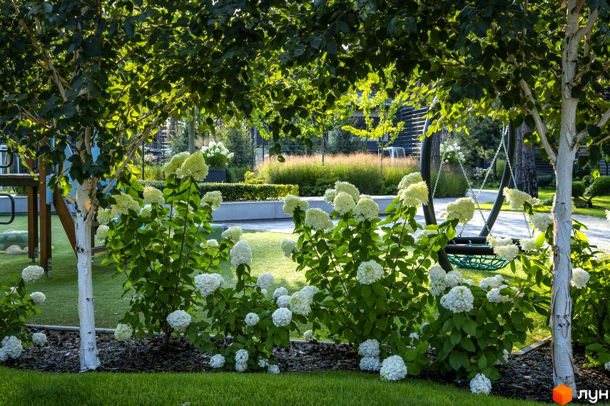 КМ ZAHRAVA village 2: зелений двір з квітучими кущами та гойдалкою, дитячий майданчик.
