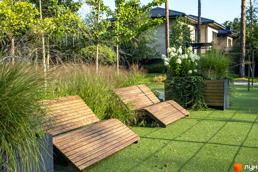 Котеджне містечко ZAHRAVA village 2 з дерев'яними шезлонгами та декоративними травами на зеленій галявині. На задньому плані видно сучасний будинок та дерева.