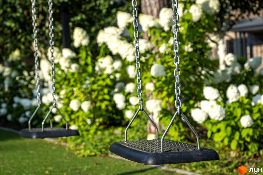 КМ ZAHRAVA village 2: дитячі гойдалки на фоні пишних кущів з білими квітами та зеленою травою.