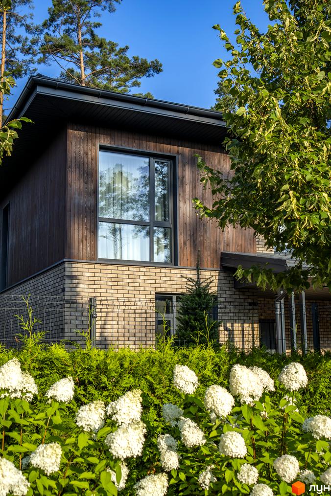 Котеджне містечко ZAHRAVA village 2. Зовнішній вигляд будинку з дерев'яним та цегляним оздобленням, великим вікном та квітучими рослинами на території.