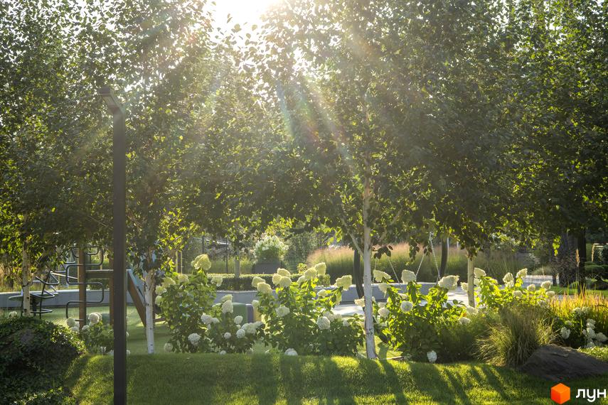 Котеджне містечко ZAHRAVA village 2 з білосніжними квітами та зеленим газоном під сонячними променями. На території розташовані дерева та елементи дитячого майданчика.