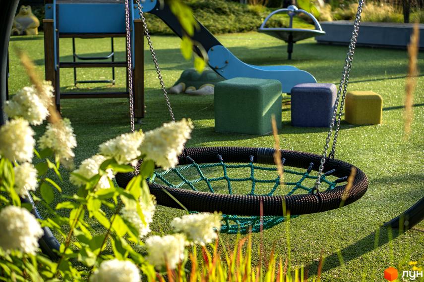 Дитячий майданчик з гойдалкою-гніздом та гіркою в КМ ZAHRAVA village 2. Зелена трава та квіти на території комплексу.