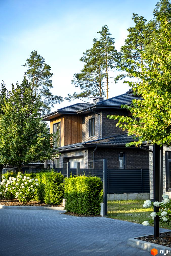 Котеджне містечко ZAHRAVA village 2. Фасад будинку з цегли та дерева, зелені насадження та квіти на прибудинковій території.