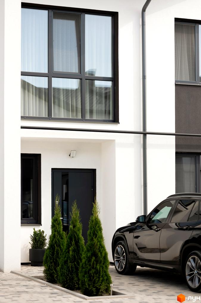 Котеджне містечко КМ WEST TOWN XIV. Фасад будинку з великими вікнами та чорними дверима. Поруч доглянута прибудинкова територія з туями та чорним автомобілем.