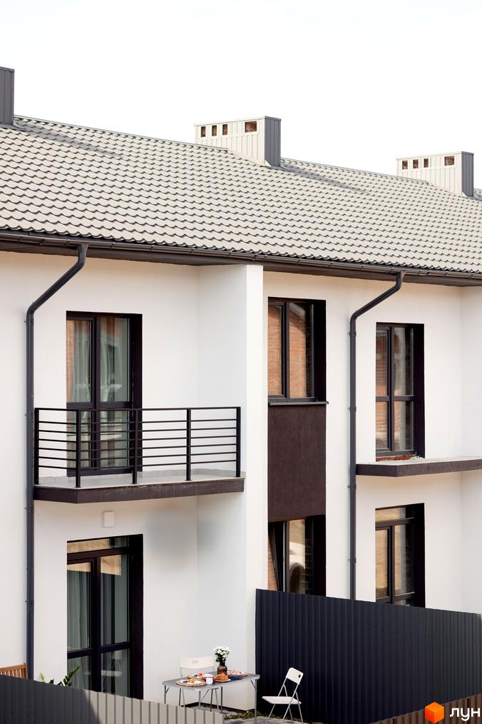 Котеджне містечко КМ WEST TOWN XIV. Зовнішній вигляд будинку з білою штукатуркою, темними вікнами та балконами, покрівлею з металочерепиці. На прибудинковій території видно паркан та вуличні меблі.