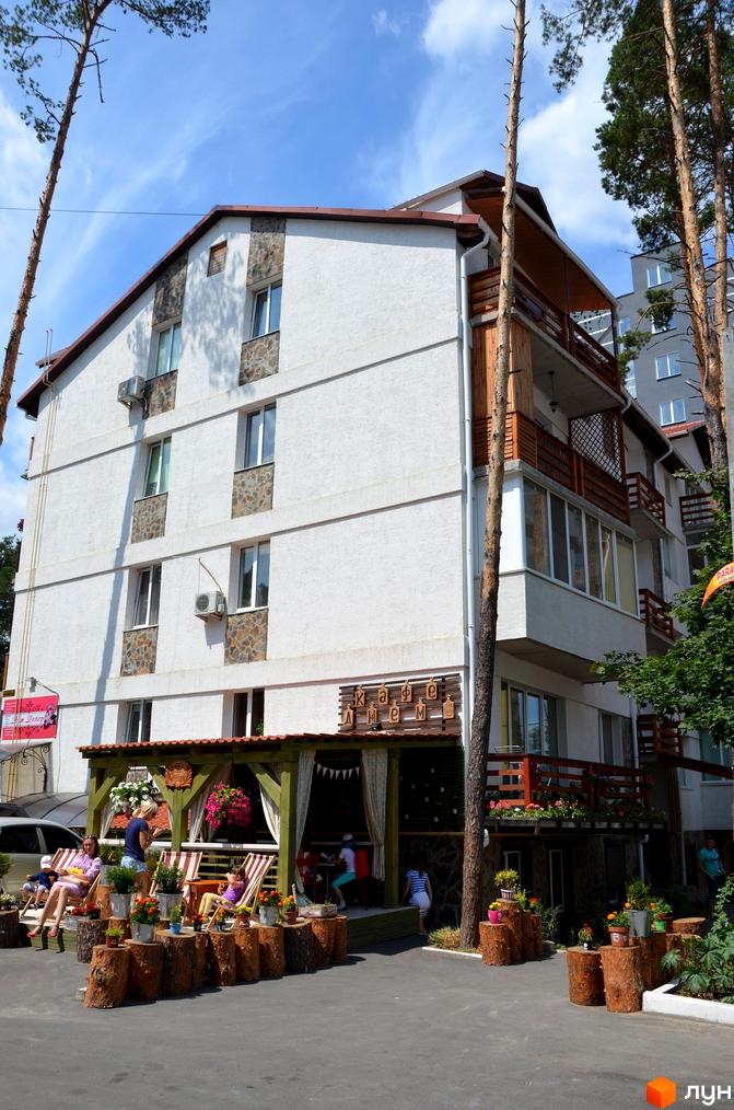 Зовнішній вигляд триповерхового житлового комплексу на вул. Університетська, 2ю, з дерев'яними елементами та балконами. Прибудинкова територія з місцями для відпочинку та зеленими насадженнями.