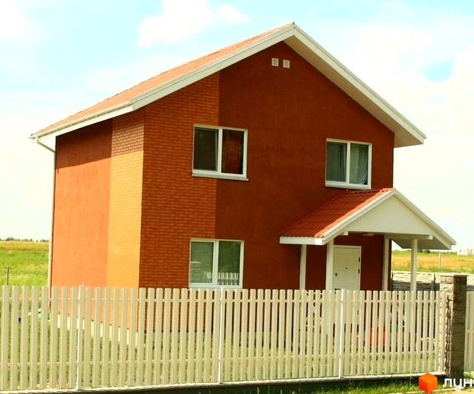 Котеджне містечко КМ Королівський сад: двоповерховий будинок з червоної цегли та штукатурки, вікна, двері, навіс та білий паркан. Навколо будинку зелений газон та поле.