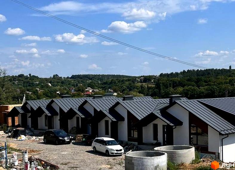 Таунхауси River Stone з сірими дахами та білими стінами на фоні зелених пагорбів. На прибудинковій території видно автомобілі та будівельні матеріали.