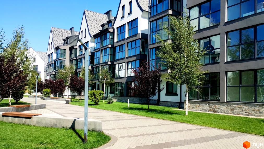 Зовнішній вигляд новобудови ЖК Desna Park Residence з великими вікнами та сірими фасадами. Поруч розташована доглянута прибудинкова територія з деревами та доріжками.