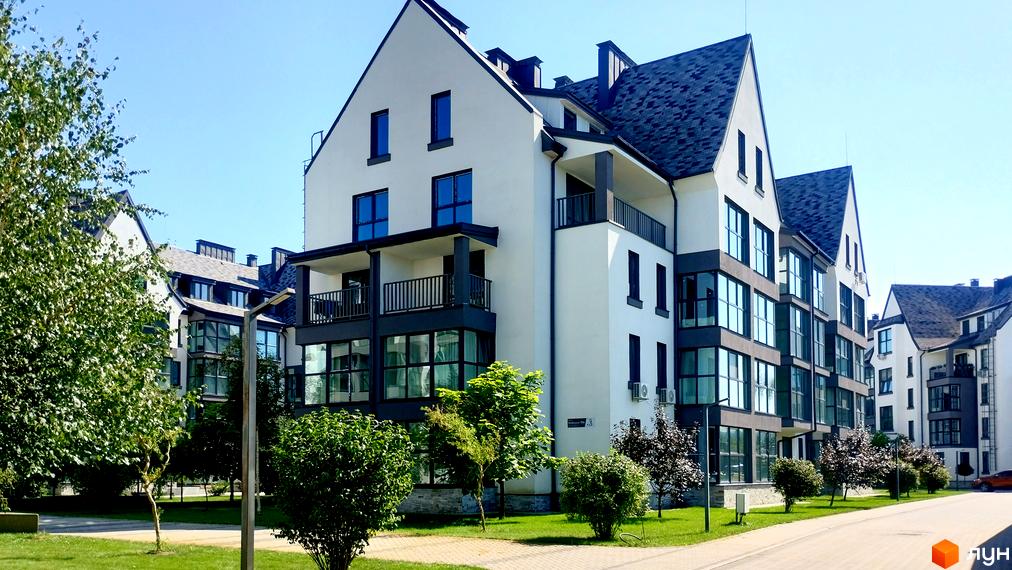 Зовнішній вигляд новобудови ЖК Desna Park Residence з білою фасадною частиною, темним дахом та великими вікнами. Навколо розташовані зелені насадження та доглянута прибудинкова територія.