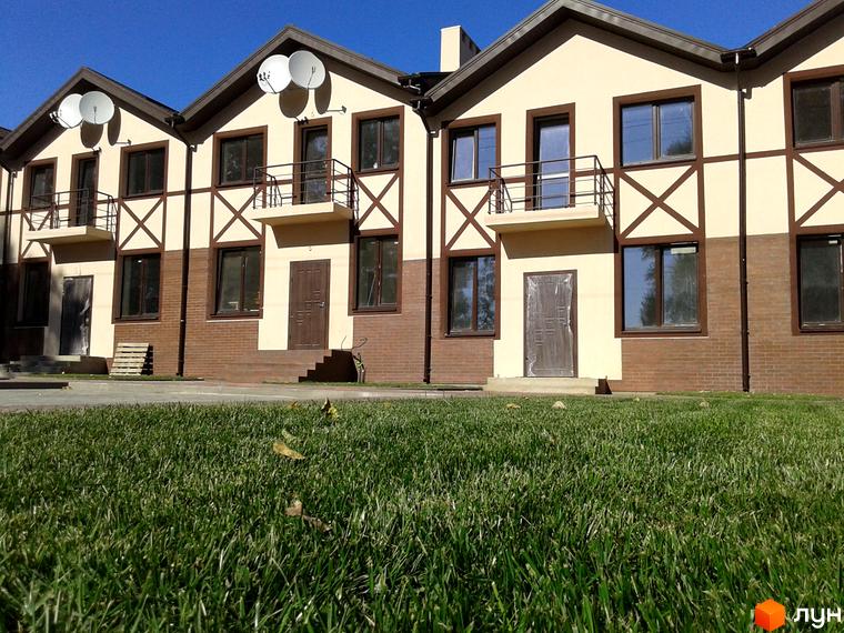 Зовнішній вигляд двоповерхових будинків з коричневими віконними рамами та балконами. Прибудинкова територія з доглянутим газоном та зеленими насадженнями.