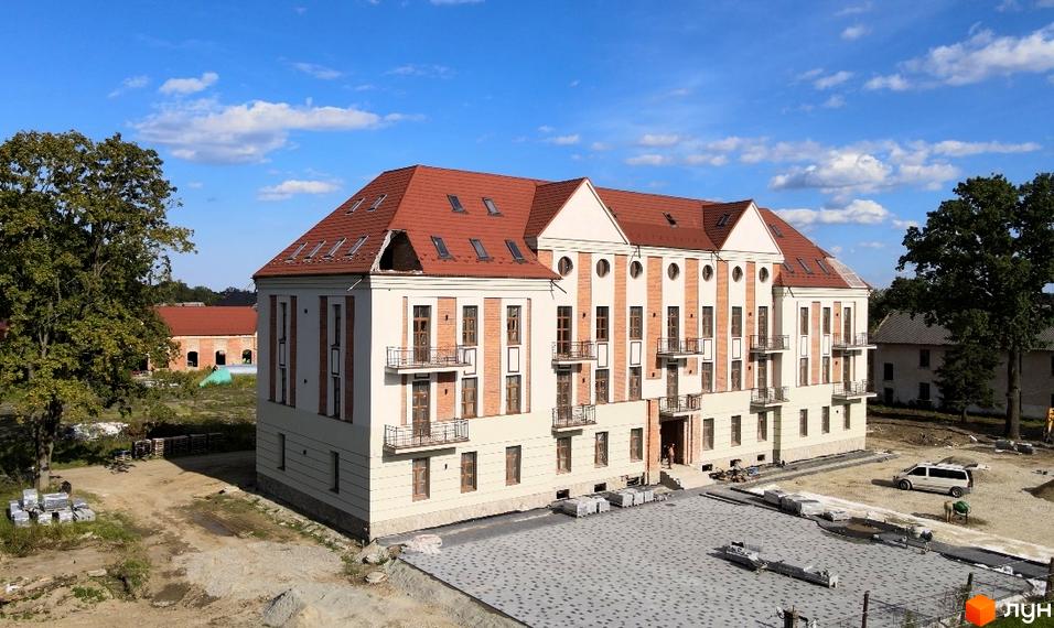 Зовнішній вигляд житлового комплексу Австрійський з червоним дахом та цегляними елементами фасаду. На прибудинковій території видно будівельні роботи та бруківку.