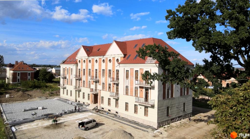 Житловий комплекс Австрійський - це чотириповерхова будівля з червоним дахом та цегляними елементами фасаду, розташована на фоні блакитного неба з хмарами. Територія навколо новобудови активно облаштовується.