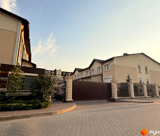 Житловий комплекс Cappuccino House - двоповерхові будинки світло-бежевого кольору з коричневим дахом, огороджені парканом з кованими елементами та автоматичними воротами. Територія перед будинками вимощена плиткою, прикрашена зеленими насадженнями.