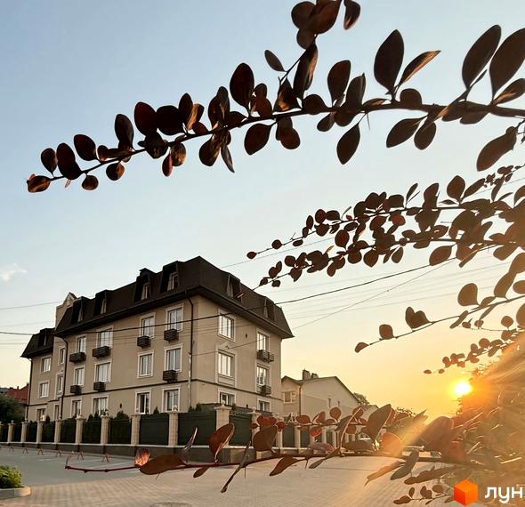 Новобудова ЖК Cappuccino House з п'ятьма поверхами та мансардою, вікнами з білими рамами та балконами. Навколо будинку розташована огорожа та зелені насадження.