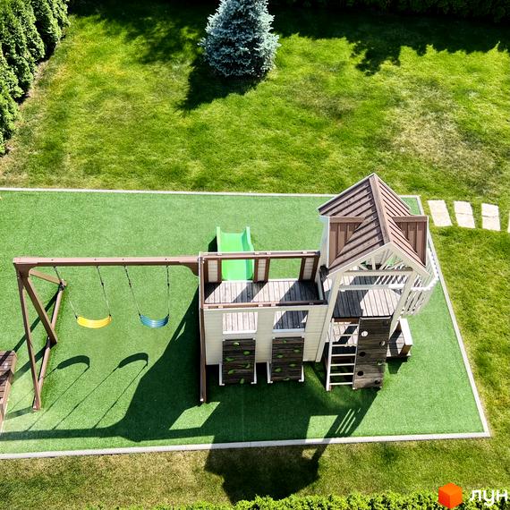 Дитячий майданчик з гіркою та гойдалками на зеленій галявині біля ЖК Cappuccino House. Навколо доглянутий газон та кущі.