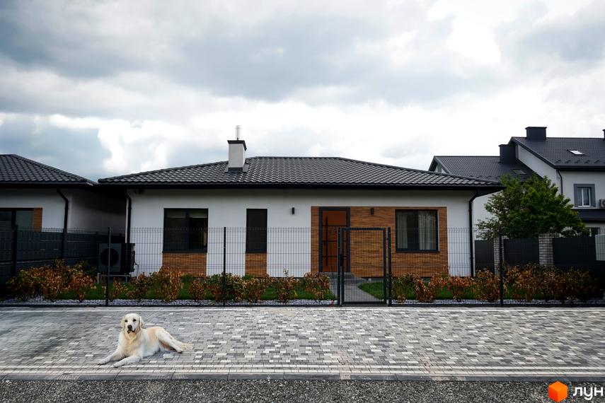 Котеджне містечко КМ MATRIX: одноповерховий будинок з білою стіною та дерев'яними елементами фасаду, огороджена територія з доглянутими кущами та вимощеною доріжкою.