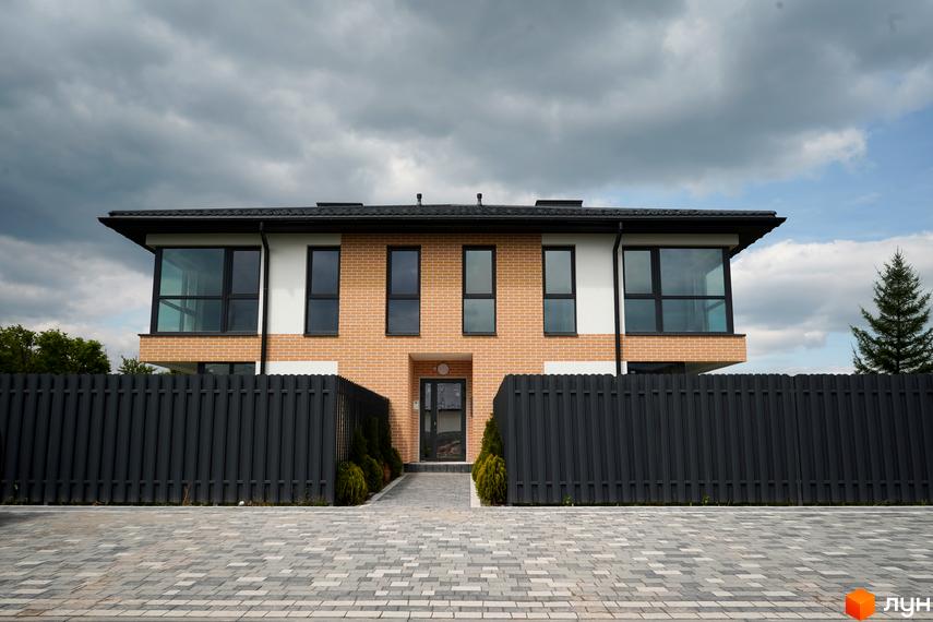 Будинок з цегляним фасадом та великими вікнами, огороджений парканом. Перед будинком вимощена бруківкою доріжка.