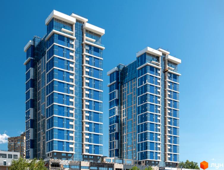 Житловий комплекс Edelweiss House - це дві висотні будівлі з блакитними вікнами та білими елементами фасаду. Навколо видно зелені дерева та блакитне небо.