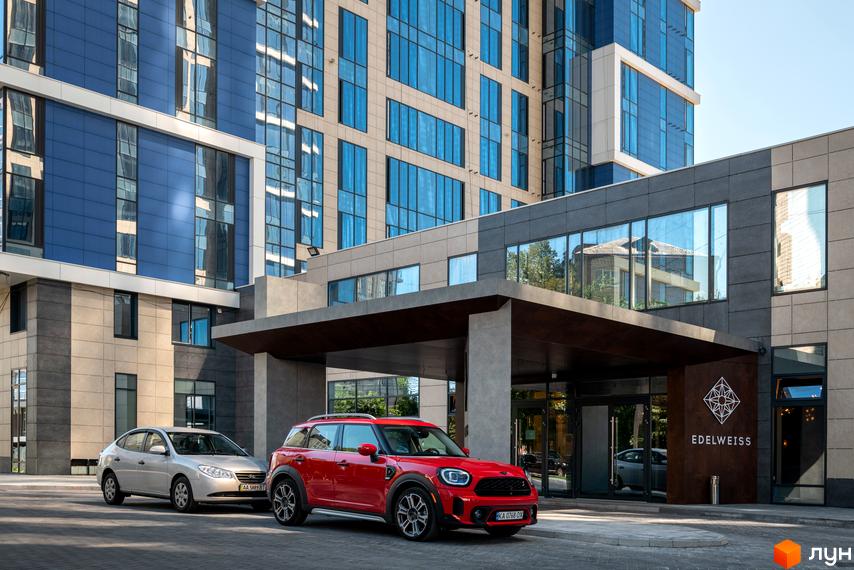 Житловий комплекс Edelweiss House з червоним та сріблястим автомобілями біля входу. Сучасна архітектура будівлі з великими вікнами.