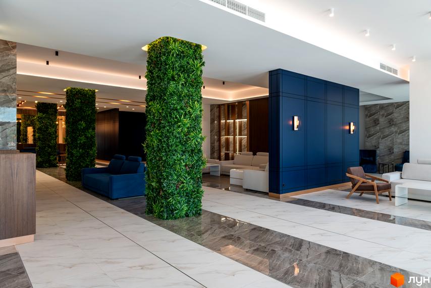Елегантний хол у житловому комплексі Edelweiss House з вертикальним озелененням та синіми акцентами. Просторий інтер'єр з комфортними зонами відпочинку.