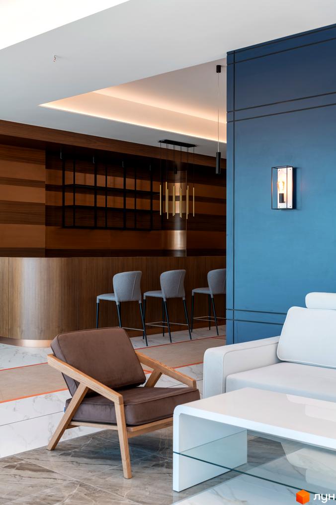 Інтер'єр вітальні з диваном та кріслом біля бару з сірими стільцями. Сучасний дизайн житлового комплексу.