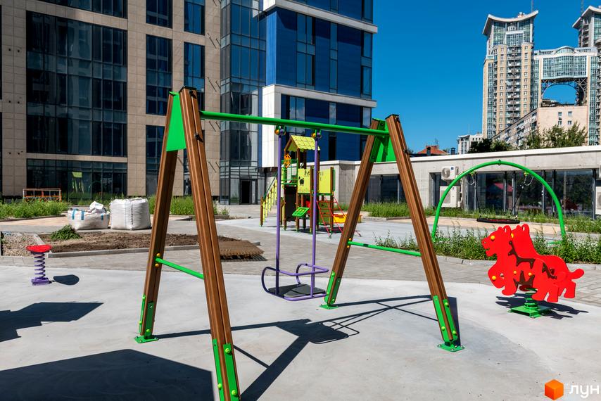 Житловий комплекс Edelweiss House з дитячим майданчиком та гойдалками. Навколо зелені насадження та сучасна забудова.