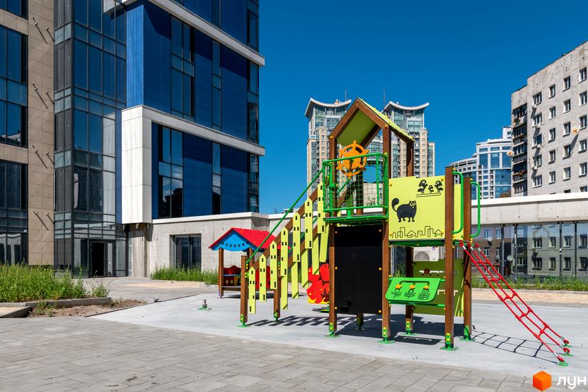 Житловий комплекс Edelweiss House з дитячим майданчиком на фоні сучасних будівель. Територія комплексу облаштована для відпочинку.