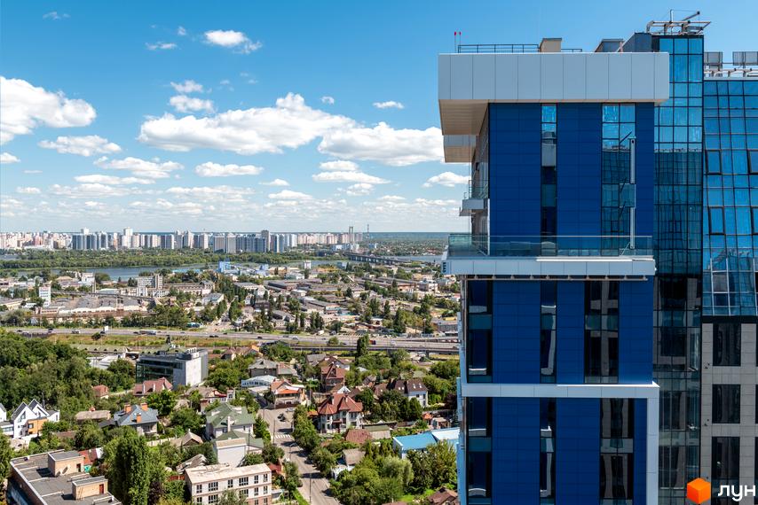 Вид на новобудову ЖК Edelweiss House з синіми скляними фасадами та білими елементами, на тлі міського пейзажу з річкою та хмарочосами.