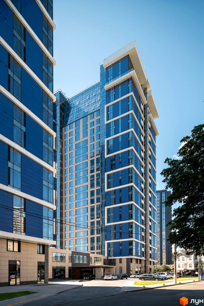 Сучасний житловий комплекс Edelweiss House з блакитними скляними фасадами та білими декоративними елементами. Територія перед будинком з доглянутим газоном та автомобілями.